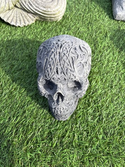 The image depicts a gray, stone skull ornament resting on a patch of artificial green grass. The skull is intricately carved with Celtic knot patterns. A portion of a gray, shell-shaped stone object is visible on the left side of the frame, also resting on the artificial grass. The scene appears to be outdoors in daylight. Although the specific location in Chirk, United Kingdom cannot be identified from the visual data, this is likely an outdoor decorative display.