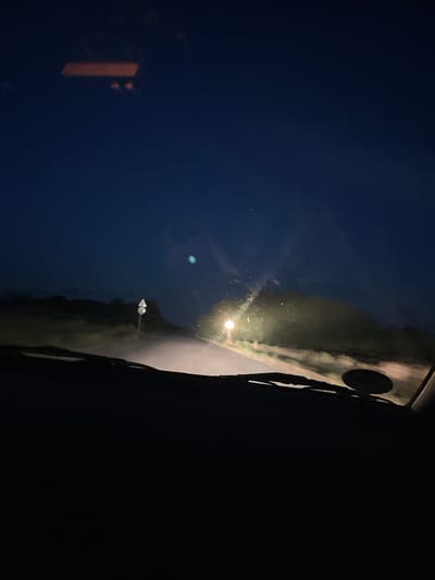 The image is a low-light, interior view from a vehicle's dashboard, looking out at a road at dusk. The sky is a deep blue, transitioning to black at the top. In the distance, a road is visible, illuminated by headlights. There is a signpost with a triangular white sign on the left side of the road. The landscape on either side of the road appears to be rural with low-lying vegetation. The windshield of the vehicle is visible in the foreground, with reflections on its surface. The dashboard itself is black and takes up the bottom portion of the image. There are no people or animals visible in the image. The location is stated to be Novo Zhelezare, Bulgaria, but this cannot be verified from the visual information in the image alone.