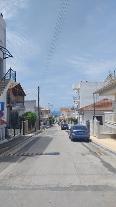 The image depicts a residential street in Νέα Μηχανιώνα, Greece, with a gentle incline and a view of the sea in the distance. The street is paved with asphalt and concrete, showing signs of wear and tear, including cracks and patches. Two cars are parked on the right side of the street, a blue sedan in the foreground and a darker sedan further up the road. The houses lining the street are typical of a Mediterranean setting, featuring white walls, tiled roofs, and balconies. Overhead power lines crisscross the sky, supported by wooden and metal poles. A speed bump is visible across the road. The sky is partly cloudy, with wispy white clouds against a pale blue backdrop. Vegetation, including small trees and bushes, is present in front of some houses. A traffic sign with blue and red elements is attached to a pole on the left side of the street. The overall scene conveys a quiet, everyday atmosphere in a coastal Greek town.