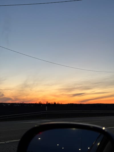 The image captures a roadside scene at twilight. On the right is a car's side mirror reflecting dim lights. Adjacent to the car, there's an asphalt road with visible markings and a barrier. Beyond the road, a line of trees is silhouetted against a vibrant sunset sky, transitioning from orange near the horizon to lighter hues higher up. On the left, the sky is a pale blue, with power lines visible. The specific location is Kuregovo, Russia, though no identifiable city elements or features are immediately apparent in the image.