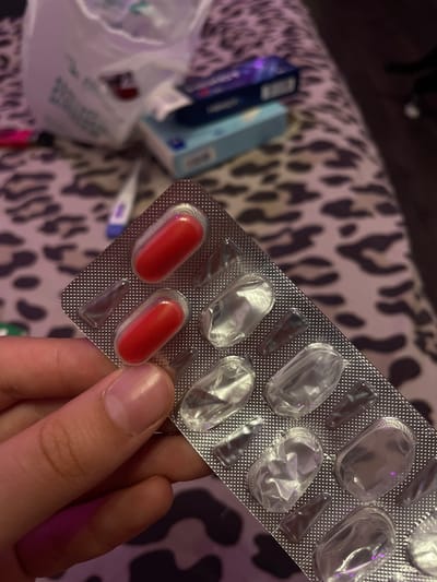 A hand holds a blister pack containing two red pills, with the remaining cavities empty. The blister pack is positioned in the foreground of the image. In the background, a leopard-print fabric is visible, serving as a surface. Behind the fabric, several items are blurred: a white plastic bag, a thermometer, and two boxes, one blue and one darker colored. The scene appears to be indoors, with artificial lighting. There is no information or indication of the location being Vaganovo, Russia.