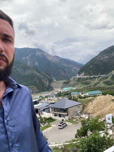 A man, partially visible, takes a selfie with a mountainous landscape in the background. He is wearing a blue, patterned shirt. The location appears to be Chalda, Russia, with a village nestled at the base of steep, green mountains. Houses with grey and green roofs are scattered throughout the village, and a road winds its way up the mountainside. The sky is overcast with clouds.