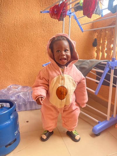 A young Black child, appearing to be between one and two years old, stands in the center of the frame, facing forward and smiling. The child is wearing a pink hooded onesie with a circular cream-colored design on the chest featuring a brown starburst pattern. The onesie has a zipper closure and is adorned with tiny buttons on the legs. The child is also wearing dark slides with a green frog design on each. The child's head is partially obscured by the hood of the onesie.
The setting appears to be an outdoor or semi-outdoor area, possibly a balcony or patio, in Iyiowa Odekpa, Nigeria. The background features a textured, orange wall with vertical ridges. To the left of the child, a large blue gas cylinder is visible, with white lettering partially obscured. Behind the child and to the right, a white clothes drying rack with blue plastic components is present, holding several items of clothing. To the left of the drying rack, stacked plastic water bottles are partially visible. The floor is tiled in a light beige color. The overall lighting suggests daylight.