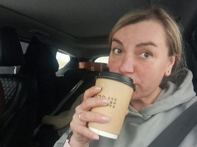 A woman in a gray hoodie and wearing a seatbelt takes a sip from a paper coffee cup with a black lid. The cup is tan and has a coffee bean design on it. The woman is inside a car, presumably the driver. Dark colored seats and a partially visible backseat passenger are also visible.