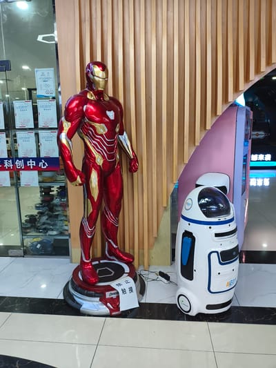 The image shows an indoor scene, likely a commercial or exhibition space, featuring a life-sized statue of Iron Man and a white robot. The Iron Man statue is in a dynamic pose, made of red and gold metallic material, standing on a circular black and red platform. A white sign with Chinese characters, meaning "Do not touch," is attached to the platform. To the right of the Iron Man statue is a white, cylindrical robot with blue accents. It has a domed head with a transparent screen displaying some graphics, and a blue panel on its side. The background consists of vertical wooden slats forming a curved wall, and a glass display case filled with various spools of colored material, possibly for 3D printing. In the distance, there are illuminated signs with Chinese characters. The floor is tiled with a marble pattern. The scene appears to be set in the city of Jiguan District, China, given the context provided.