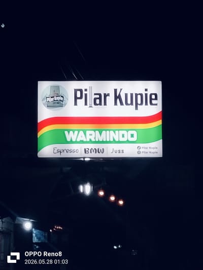 At night, a sign illuminated above a shop or building reads "Pilar Kupie" with a logo of a building. Below the name is "WARMINDO" and "Espresso BMW Juss". There is a green, yellow and red stripe between the store name and warmindo. The image was taken with an OPPO Reno8, indicated by the watermark in the lower left corner, time-stamped 2026.05.28 01:03. The location is likely Cempeudak, Indonesia, based on the implied context of the brand name. The background is dark and out of focus, with indistinct street lights.