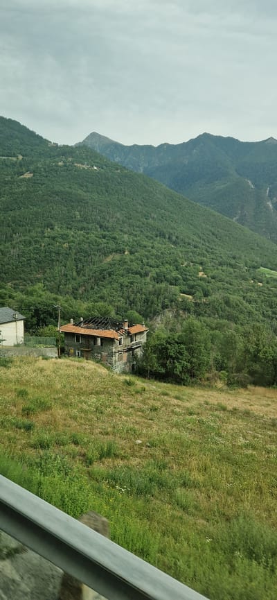 An aerial view shows a stone house with a damaged roof, likely from a fire, situated on a grassy hill in Challand-Saint-Victor, Italy. The house is a two-story structure with an orange tiled roof, most of which appears to be missing, exposing blackened beams underneath. Several windows are visible, some of which appear broken. The house is surrounded by overgrown grass and vegetation, with a few trees in the background. In the distance, lush green mountains dominate the landscape, with a cloudy sky overhead. To the left of the house, a portion of another building is visible, appearing to be a separate, intact structure. A grey metal railing runs along the bottom of the image, suggesting the photo was taken from a vehicle or elevated viewpoint.