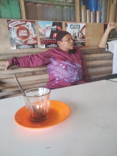 A glass of liquid with a spoon sits on an orange saucer on a white table. A man wearing a burgundy shirt and a Muslim cap is sitting in the background. Behind him are posters, including "Lucky Strike," "Dunhill," and "CLU13." A second man in a white shirt is partially visible with his arm raised. The location is Pulorayeuk, Indonesia.