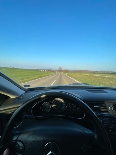 The image presents a first-person perspective from within a moving Mercedes-Benz vehicle, traveling on a rural asphalt road under a clear, bright blue sky in the vicinity of Weißenhorn, Germany. The time appears to be late afternoon or early evening, indicated by the low sun angle and soft lighting.
In the immediate foreground, the black leather steering wheel, emblazoned with the Mercedes-Benz logo, occupies the lower central portion of the frame. A driver's left hand is partially visible gripping the lower left section of the steering wheel. Behind the wheel, the vehicle's instrument cluster is visible, showing a speedometer needle resting near 90 km/h, a digital readout confirming 91.0 km/h, an external temperature of 23.0°C, and a remaining fuel range of 260 km. The dashboard is dark-colored, and air vents are visible on the passenger side. The windshield displays some minor dust or debris.
The two-lane road stretches straight ahead, marked by solid white lines on the shoulders and a dashed white center line, eventually curving slightly to the right in the distance. Lush green fields flank both sides of the roadway. On the left side, a wooden hunting blind or deer stand is visible in the field, with a small cluster of houses, indicating a hamlet or village, further on the horizon. The right side features additional green fields bordered by a line of leafless deciduous trees, suggesting a late autumn or winter season. A solitary, bare tree stands prominently near the road's vanishing point in the background. No other vehicles or individuals are discernibly present on the road.