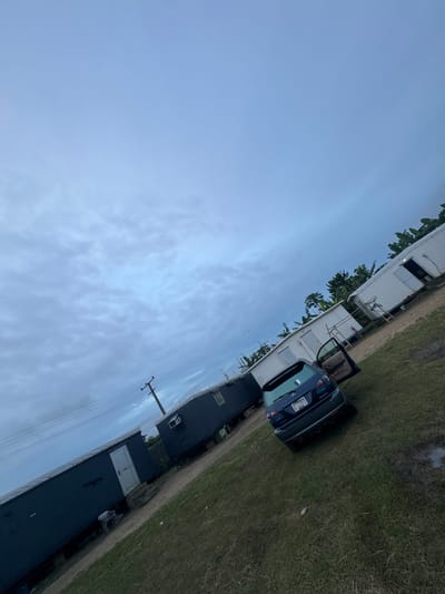 The image shows a scene in Igwuruta, Nigeria. A blue hatchback car with its driver-side door open is parked on a grassy verge next to a white mobile home or trailer. The surrounding environment includes more structures, potentially residential, some with dark roofs. A utility pole with power lines is visible. The sky is cloudy and pale blue. There are no people visible in the image.