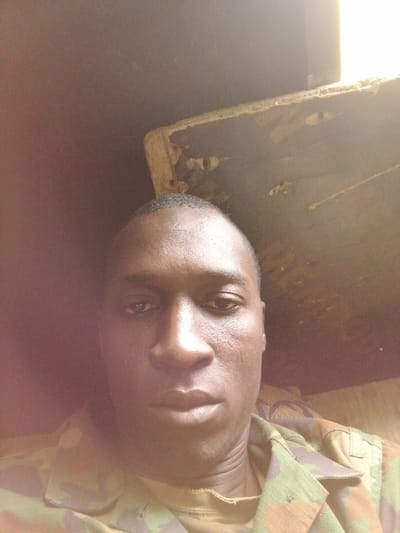 The image displays a close-up, chest-up portrait of an adult male. The subject has dark skin, short, closely cropped dark hair, and a neutral expression, looking directly towards the camera. He is wearing a camouflage uniform shirt, visible from the neck to the mid-chest, featuring a pattern of green, brown, and dark hues. The background, located in Kayauki, Nigeria, shows an indoor or enclosed environment. In the upper right quadrant, a section of a light-colored, distressed wooden or particle board structure is visible, exhibiting significant wear and chipping along its edge. A bright light source is positioned behind this structure, creating an intense highlight on its perimeter. The remainder of the background is a uniformly dark brown/black, indicating a dimly lit space. No specific actions or interactions are depicted.
