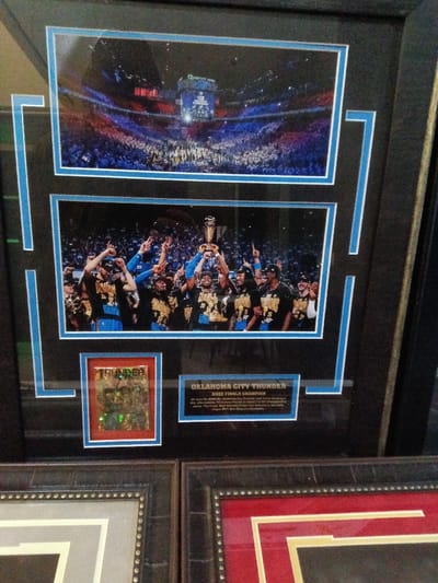 Framed memorabilia showcases the Oklahoma City Thunder's 2025 NBA Finals Championship. Above, a wide shot of the arena during the finals. Below, a photo of the team celebrating with the trophy. A small, framed emblem with "THUNDER" in stylized font is included, along with a descriptive plaque stating "OKLAHOMA CITY THUNDER 2025 FINALS CHAMPION." The item is professionally framed with a black frame and a blue inner border.