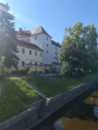 The image depicts a view of the Otava River in Písek, Czechia. On the left, a historic building with a sign reading "RESTAURACE" is visible, its yellow facade contrasting with the white upper stories and tiled roof. A lush green lawn slopes down to the river, dotted with steps leading up to the restaurant's terrace, where tables and chairs are set. Several people are seated on a stone wall overlooking the river. Large, mature trees with dense green foliage flank the building and the riverbank. The river itself is dark and reflects the surrounding greenery and the sky. A stone embankment lines the river on the right side. The scene is bathed in sunlight, casting shadows and highlighting the vibrant colors of the environment.
