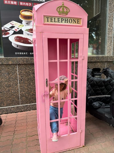 A young girl wearing a pink cap, a light pink t-shirt with a cartoon image, and blue pants stands inside a bright pink, vintage-style telephone booth. She is gripping the bars of the booth with her right hand and holding the brim of her cap with her left. The booth has a gold-colored "TELEPHONE" sign with a crown emblem at the top. The ground outside the booth is paved with red, interlocking bricks. To the left of the booth is a large black poster featuring images of Chinese cuisine, including Peking duck. Behind the booth on the right is a dark-colored, quilted motorcycle cover. The background shows a building with a granite-like facade and a window. The scene appears to be on a street in Hunchun, China.