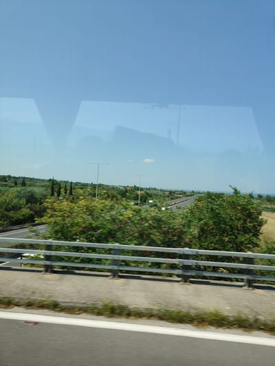 The image depicts a view from a moving vehicle on a sunny day, likely along a highway in the vicinity of Πλάκα Λιτοχώρου, Greece. The foreground shows a concrete barrier and a road with white lines. A metal guardrail runs along the edge of the highway. Beyond the guardrail, lush green trees and bushes are visible, with some showing motion blur due to the vehicle's movement. In the background, a multi-lane highway stretches into the distance, with some vehicles present. Cypress trees and other vegetation are scattered across the landscape, and hints of buildings can be seen in the far distance. The sky is a clear, pale blue, with a faint reflection of what appears to be a vehicle's interior and text on a window, possibly indicating "ISSUE DE SECOURS" (emergency exit), suggesting the photograph was taken through a window. The scene is characterized by natural scenery and infrastructure typical of a highway environment in Greece.