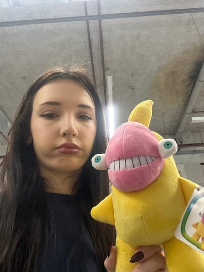 A close-up, eye-level shot captures a young woman holding a distinctive yellow plush toy. The image appears to be taken as a selfie, with the woman looking directly at the camera with a neutral expression.
The woman occupies the lower portion of the frame. She has long, dark brown hair that falls over her shoulders and part of her chest. Her skin is fair, and she has light makeup, including defined eyebrows, subtle eyeliner, and dark pink or reddish-brown lipstick. She is wearing a dark-colored top, possibly black or dark blue, visible below her chin.
To the right and slightly above the woman's face, a bright yellow plush toy is held up by her hand. The toy features a large, cartoonish pink mouth with prominent white teeth that are outlined in black, giving it a wide, toothy grin. Two large, googly eyes with green irises and black pupils extend from the top and bottom of the pink mouth. The toy also has small, yellow fin-like protrusions on its body. A white tag with colorful graphics (including hints of green, orange, and blue) is visible attached to the top of the toy, near the woman's hand. The woman's hand is visible on the right, holding the yellow toy, and her fingernails are painted with a dark red or maroon polish.
The background is an indoor setting with an industrial or utilitarian appearance. It consists of a grey, concrete-like wall or ceiling with visible pipes and light fixtures. There are horizontal dark lines, possibly support beams or conduits, running across the concrete surface. Patches of darker discoloration or stains are visible on the grey surface in the upper left portion of the image. The lighting appears to be artificial, likely from overhead fixtures, casting a somewhat cool tone on the scene.