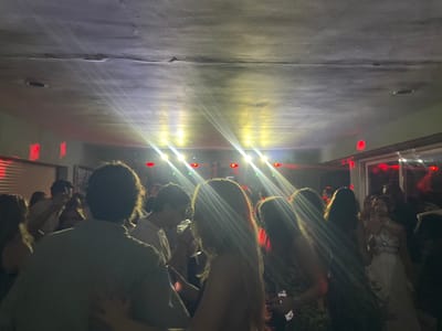 The image depicts an indoor scene with a gathering of people, likely at a party or event. The lighting is dim and atmospheric, with bright spotlights creating lens flares across the upper portion of the image. Red lights are visible on the walls in the background. A group of individuals, mostly young adults, are gathered in the space, with some facing the camera and others engaged in conversation or dancing. The ceiling appears to be textured and dark. The environment suggests a social gathering taking place in a venue. While the specific location in Pozuelo de Alarcón, Spain, is not discernible from the visual elements, the overall impression is that of a lively indoor event.
