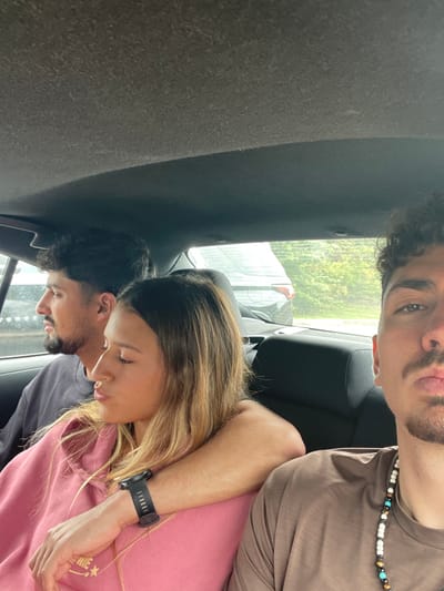A photograph taken within the rear passenger compartment of a vehicle depicts three individuals. The vehicle interior has a dark gray fabric headliner and dark-colored seats. On the far left, a male with dark hair and a beard, wearing a dark shirt, is seated, gazing out the left window. In the center, a female with long, light brown hair, dressed in a pink hooded sweatshirt and wearing a black wristband, is positioned with her eyes closed or looking downwards and lips slightly pursed. Her left arm is draped over what appears to be the shoulder or seatback of the adjacent passenger. On the right, a male, partially visible from the nose down, with dark, curly hair and a beard, wearing a brown t-shirt and a beaded necklace, faces the camera with pursed lips, indicating he is likely taking the photograph. Through the rear window, another vehicle, a light-colored SUV, is visible behind the depicted car, followed by green trees. The specific location within the city of Oxon Hill, United States, cannot be determined from the image.