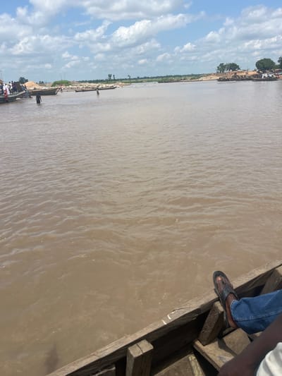 A wide, muddy river flows through a scene of human activity in Nemnei, Nigeria. Several wooden boats are visible, some carrying passengers and others appearing to be used for transport of goods, possibly sand or gravel, given the piles on the riverbank. People are seen wading in the shallow water near the bank, and some are standing on the boats. The riverbank is lined with sand deposits and sparse vegetation, with trees and buildings visible in the distance. The sky is partly cloudy with white clouds against a blue backdrop. In the foreground, the wooden interior of a boat is visible, with a person's legs and feet, wearing blue jeans and black sandals, resting on the edge. The water appears calm with small ripples.
