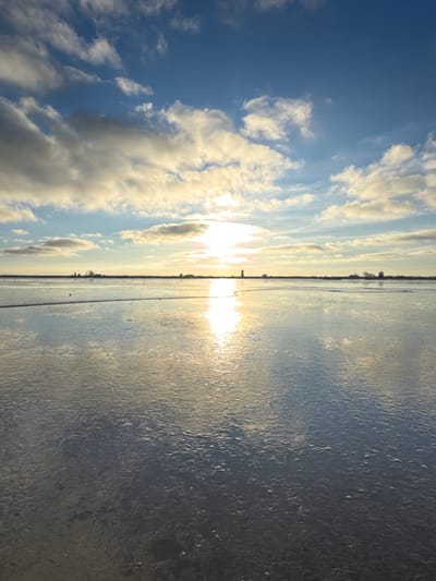 A wide-angle, low-elevation image depicts a flat, reflective landscape under a partly cloudy sky, characteristic of the Broadland district, United Kingdom. The sun, positioned low above the distant horizon slightly right of center, emits a strong glare that creates a prominent vertical reflection across the foreground. The sky is bright blue with scattered white and light grey clouds. Along the distant, linear horizon, a dark, cylindrical tower structure is discernible to the right of the sun's reflection, alongside several other indistinct distant buildings or tree clusters. The expansive foreground consists of a calm, reflective surface, consistent with shallow water, wet mudflats, or a sheet of ice, which mirrors the blue sky and solar reflection. The surface exhibits minor textural irregularities but no significant water movement or distinct objects within the immediate foreground. No individuals, animals, or explicit actions are present.