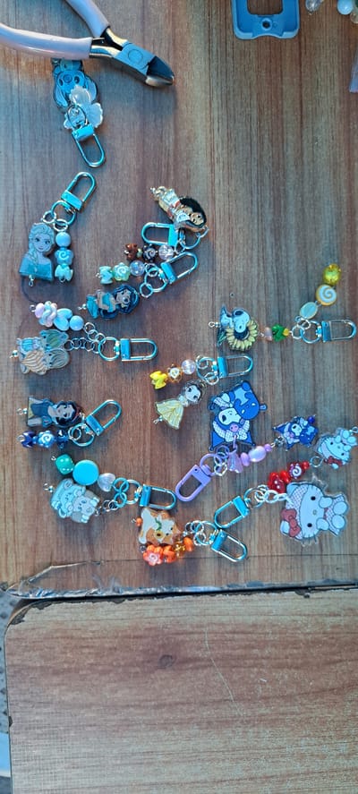 The image shows a collection of handmade keychains laid out on a wooden surface. The keychains feature various charms, including Disney princess characters like Elsa, Jasmine, and Snow White, as well as Snoopy, a blue alien creature, and Hello Kitty. Some keychains are also adorned with colorful beads and small flower charms. A pair of pink-handled wire cutters is visible in the upper left corner of the image. The scene is illuminated by overhead lighting, casting soft shadows on the wooden surface. The keychains appear to be arranged for display or for sale. The overall impression is one of a craft or hobby-related setting. The specific location in the city of Mandaluyong, Philippines, is not determinable from the image.