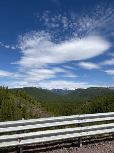 Anavgai, Russia - A scenic vista overlooks a vast expanse of rolling, tree-covered mountains under a partly cloudy blue sky. In the foreground, a silver metal guardrail with three horizontal bars runs across the bottom of the frame, separating the viewer from the mountainous landscape. The guardrail is attached to vertical metal posts, and a small red reflector is visible on the bottom bar. The ground in front of the guardrail consists of dark, gravelly soil. The mountains in the distance are covered in dense green coniferous forest, with some areas of rocky, barren slopes. In the far distance, the peaks of some mountains appear to have patches of snow. The sky is a vibrant blue, dotted with scattered white cumulus clouds, some of which are elongated and wispy.