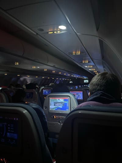 The image depicts the interior of an airplane cabin at night. Overhead compartments are visible, along with illuminated signs indicating "Fasten Seatbelt" and "No Smoking." Several rows of passenger seats with individual entertainment screens are visible. Some screens display flight path maps, while others are dark. Passengers are seated in some of the chairs, though their faces are largely obscured by the dim lighting and their positions. One passenger in the foreground is wearing a dark hoodie and a purple garment. The overall atmosphere is dark and confined, typical of an airplane cabin during a night flight. The location is specified as Sisa Chorakhe Noi Subdistrict, Thailand, however, the visual information in the image does not provide any specific cues to confirm or deny this location.