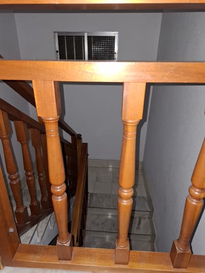 A ground-level, medium-shot interior view shows a wooden staircase viewed through the wooden balustrade. The stair treads are visible, with decorative turned balusters. A window with a security grille is visible in the background to the left. The walls are painted gray. The ceiling is white. The top of the stairs, tiled in white marble, is visible. There is no evidence of people in the photo. The location is unknown.