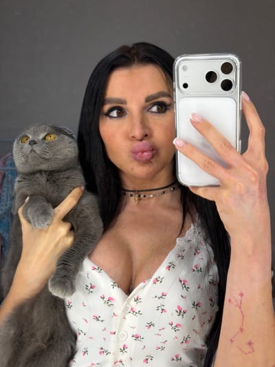 An adult female, identified as being in Svkh Voronezhskiy, Russia, is depicted in an indoor setting with a plain dark grey background. She has long dark hair, prominent eyebrows, and significant eye makeup. Her lips are pursed in a "kissing" expression, and her gaze is directed towards the smartphone she holds in her left hand, indicating she is taking a selfie. The phone is light-colored, housed in a clear case, and features a triple-lens camera array on its rear. Her fingernails display a French manicure. She wears a white V-neck top with pink floral patterns and lace trim, revealing cleavage. A black choker necklace with beads and a small golden cross pendant is visible around her neck. On her left forearm, a faded pink tattoo resembling a constellation is present. She cradles a grey Scottish Fold cat in her right arm. The cat has amber eyes and is looking upwards and to its right.