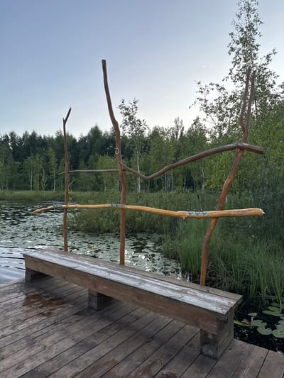 A rustic wooden bench sits on a wooden dock next to a calm body of water covered in lily pads. A structure made of natural branches, possibly for hanging items or as decoration, stands behind the bench. The background is filled with lush green trees and foliage, suggesting a natural, possibly park-like setting. The sky is clear and pale blue, indicating either early morning or late evening. The location is identified as Pchelka, Russia.