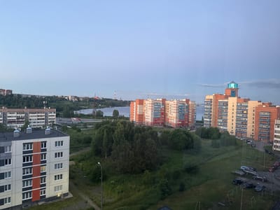 This image shows a city skyline view at dusk in Kondopoga, Russia. In the foreground, a white and gray apartment building with orange and dark gray accents stands prominently. Beyond this, more apartment buildings of varying heights and colors are visible, with the tallest featuring a distinctive blue glass structure on its roof. A body of water, possibly a lake or river, stretches across the middle ground, with distant industrial structures and more buildings on the opposite shore. The foreground and middle ground are filled with green trees and grassy areas, interspersed with roads and parked cars. A streetlamp is visible in the lower-middle section of the image. The sky is a soft, gradient blue, indicating the transition from day to night. There are no people visible in the scene.