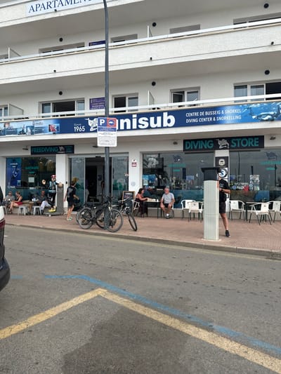 The image displays a street-level view of a white multi-story building in L'estartit, Spain. The ground floor accommodates a commercial establishment, "UNISUB Diving Center & Snorkelling," and a "DIVING STORE," indicated by blue and green signage. The building's upper floors feature balconies and an "APARTAMENTOS" sign, with a "VACATIONAL RENTALS" sign visible on a second-story balcony.

In the foreground, an asphalt road displays yellow and blue painted lines delineating a parking area. A partial view of a dark grey vehicle bumper is visible on the far left. A tall, dark grey street light pole stands centrally on the sidewalk, affixed with a blue "P" parking sign.

Approximately ten individuals are present on the sidewalk. To the left, near the "FREEDIVING" section of UNISUB, a mannequin dressed in a red helmet, black wetsuit, and flippers, holding a spear gun, stands on a yellow platform. Adjacent to the mannequin, three individuals are seated at an outdoor wooden bench and table. A person in dark clothing walks towards the UNISUB entrance. Two bicycles, a black mountain bike and another dark-colored bicycle, are parked on the sidewalk near the street light pole. Further to the right, two individuals, a woman in a dark shirt and a man in a grey t-shirt, are seated at an outdoor table with white chairs. A man in a black t-shirt and shorts stands near a white pillar on the right side of the sidewalk, facing towards the right. The large windows of the diving center display diving equipment and PADI logos.