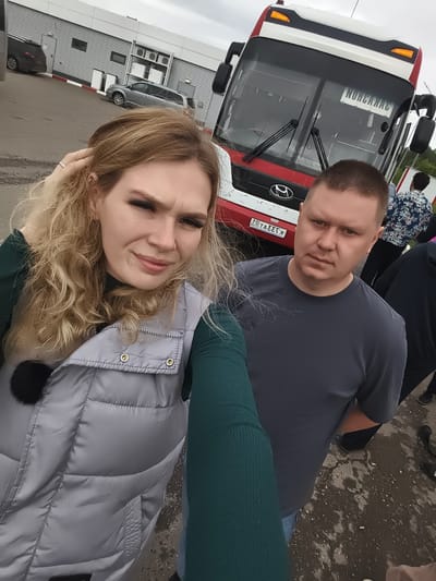 A young blonde woman is taking a selfie in a paved outdoor area in Можга, Russia. She wears a dark green long-sleeved top and a light gray quilted vest with a black faux fur pom-pom. Her right hand, adorned with a ring, touches her hair. Her expression is a slight smirk, looking directly at the camera. A man with short hair and a dark gray long-sleeved t-shirt stands to her right, looking towards the camera with a neutral expression. A silver chain is visible around his neck.
In the background, a large red and white Hyundai passenger bus is prominently featured. Its front displays a mirrored Russian license plate, "КЭЭ740Р", which would be "Р047ЭЭК" when unmirrored. A mirrored word, "НОНСАЙАС", is visible on the destination sign in the windshield. Behind the bus and people, an industrial or commercial building with corrugated metal siding, windows, and mounted air conditioning units is visible. Two parked cars, one dark and one light gray, are also in the background. The ground is paved, appearing damp or wet in patches, with a red and white striped curb marking. An out-of-focus individual in a patterned shirt is visible behind the man, facing away. The sky is overcast.