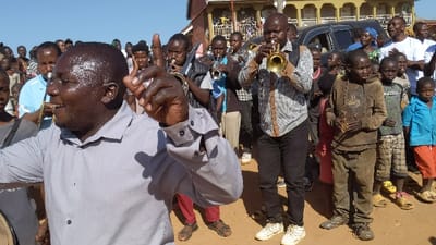 A group of people are standing in an open-air area in Kayna, Democratic Republic of the Congo. In the center of the scene is a man wearing a grey shirt who is raising his right hand as he plays a trumpet. He is surrounded by other musicians, some playing trumpets, others holding drums. Some people are watching the musicians play, while others are standing behind them. There is a black car parked behind the musicians. The ground is dusty, and the sky is clear blue. The overall scene is that of a celebration.  The exact location of the image, based on the coordinates provided, is at -0.609 latitude and 29.173 longitude in the city of Kayna, Democratic Republic of the Congo.