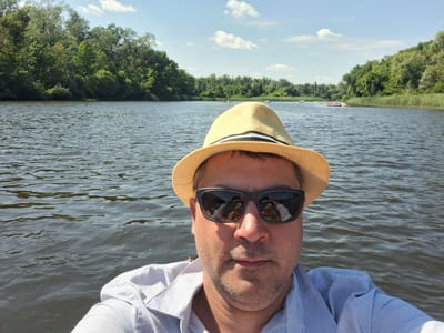 A selfie shows a man wearing a straw hat and sunglasses, with the reflection of a boat and trees visible in his sunglasses. He is in a body of water with a treeline and reeds along the shore. Several other boats, some with people in them, are visible in the distance on the water. The sky is blue with scattered clouds. The location is the river in Potoky, Ukraine.