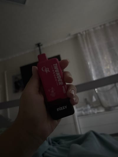 A hand is holding a pink disposable vape that says "THUNDER 8500" in white lettering. The vape also has a graphic of what appears to be a lightning bolt. The background is dimly lit and appears to be an indoor setting with a curtain visible. There is no identifiable information to place this image in Заплавская, Russia.
