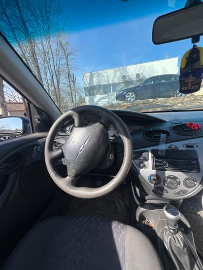 The scene is viewed from the interior of a Ford vehicle, likely a Focus generation 1 or 2, looking forward through the windshield. The driver's perspective shows a black, worn steering wheel with a "Ford" emblem and "AIRBAG" inscription. To the right of the steering wheel, the dashboard includes a car radio, climate controls, and air vents, one of which has a red "I love you" sticker. A manual transmission gear shifter is visible to the right of the driver's seat, which is upholstered in dark fabric with a subtle patterned design. A black rubber floor mat is present. A yellow and black "SPORT" branded air freshener with a ship design hangs from the rearview mirror. The left side mirror is also visible.
Outside the vehicle, a clear blue sky dominates the upper portion of the frame. Bare deciduous trees stand to the left and center. In the midground, a modern, two-story white building with a horizontal gray stripe is visible. Two external air conditioning units are mounted on its facade. A dark blue/black sedan, consistent with an Audi A4 (B7 generation), is parked on an elevated dirt mound in front of the white building. To the far left, a portion of an older structure with a reddish-brown tile roof is visible. The location is in Venets, Bulgaria. No people are visible.
