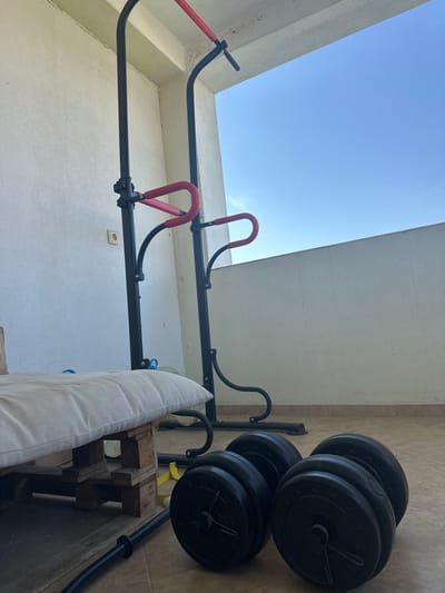An outdoor home gym setup is depicted on a tiled balcony or terrace in Marikostinovo, Bulgaria. The scene features a clear, bright blue sky visible above a low, light-colored partition wall on the right. The flooring consists of light brown square tiles. A black metal power tower with red padded grips for pull-ups and dips stands centrally. To the right of the power tower, two black adjustable dumbbells are positioned on the floor. A black barbell bar is partially visible on the floor in the lower-left foreground. A makeshift bench or seat is present in the lower-left, constructed from a white, tufted cushion resting on a wooden pallet. A yellow strap and a blue object are partially visible on the floor near the pallet. The surrounding walls are a textured off-white or cream color, with a light brown electrical outlet visible on the left wall. No individuals are present.