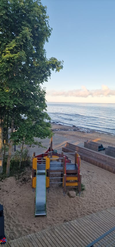 The image depicts a children's playground situated on a sandy beach next to a body of water, likely the Baltic Sea, given its location in Vārzas, Latvia. The playground features a colorful structure with yellow and red accents, including a green slide. To the left of the playground, a large tree with dense green foliage dominates the scene, casting a shadow on the sand. In the background, the sea stretches to the horizon, with a few wispy clouds visible in the pale blue sky. The beach itself is a mix of sand and small stones. Wooden decking is visible in the foreground and to the right of the playground, suggesting a boardwalk or seating area. The scene appears to be set during the daytime, possibly early morning or late afternoon, given the soft lighting. There are no people visible in the image.