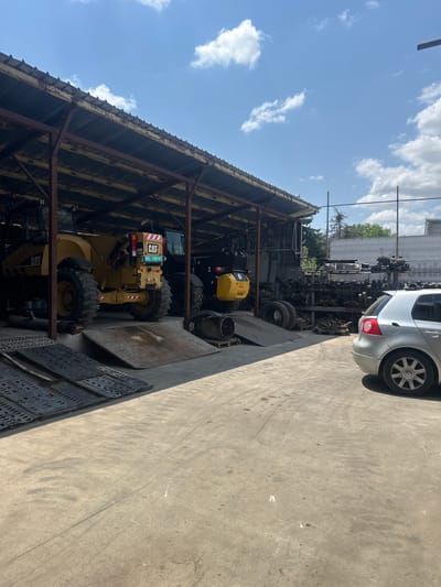 The image depicts an outdoor scene at a junkyard or auto repair facility. The dominant subjects are several large pieces of heavy machinery, including yellow Caterpillar construction vehicles, parked under a rustic, open-sided metal-roofed shelter. In the background, there are rows of assorted car parts and another silver car. The ground is paved with concrete.
The time of day appears to be daytime, with bright sunlight casting shadows. The sky is blue with scattered white clouds, suggesting fair weather. The location is identified as Hotantsa, Bulgaria. There is no discernible activity or event taking place; the scene is static, suggesting a storage or repair area.
Notable details include the branding "CAT" on the construction equipment and a license plate on one of the vehicles that reads "MD14PWX". The overall impression is one of a functional, utilitarian space where vehicles and machinery are stored or dismantled. There are no people visible in the image, and the mood is neutral, focusing on the objects and the environment.