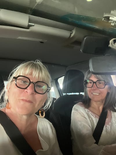 Two women wearing seatbelts are in a car. The woman in the foreground has short blonde hair and black glasses. The woman in the background has grey hair and black glasses and is smiling. The interior of the car is visible, including the rearview mirror and passenger-side sun visor. The photo was not taken in Palavas-les-Flots, France.