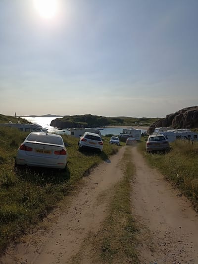 The image depicts a dirt road leading through a grassy, coastal landscape with several cars and caravans parked on either side. In the background, a body of water with islands and a rocky shoreline is visible under a bright sky. The location is identified as Carnboy, Ireland. There are no people visible in the scene. The cars and caravans suggest a campsite or parking area. The environment is natural, with vegetation and water.