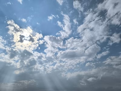 A wide-angle view of the sky above Chamanzar, Uzbekistan, is presented. The scene is dominated by a partially cloudy sky, featuring a mixture of bright white and light to dark grey cumulus and stratocumulus clouds. The sun, partially obscured by clouds in the upper-left quadrant, emits visible crepuscular rays that penetrate through gaps in the lower cloud cover, extending diagonally downwards towards the lower-left and lower-right portions of the frame. Patches of clear blue sky are visible between the cloud formations. No ground features, structures, people, or man-made objects are discernible within the frame.