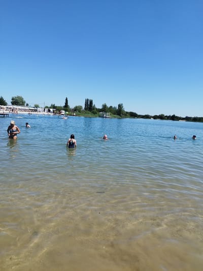This media file depicts a sunny day at an outdoor swimming area. The scene features a clear blue sky and calm, shallow water. Several people are visible in the water, enjoying a swim. In the foreground, a woman in a patterned bikini stands in the water, while further out, a young girl in a dark swimsuit wades. Other individuals are scattered throughout the water, some appearing to swim or float.

The background shows a sandy beach area with white cabanas or umbrellas providing shade. Trees line the far shore, and some buildings or structures are visible in the distance. The overall atmosphere appears relaxed and leisurely, suggesting a recreational outing on a warm day. The time of day is likely midday or early afternoon due to the bright sunlight. The location cues, such as the type of beach setup and vegetation, are consistent with a summer resort or public swimming area. There is no visible text in the image.