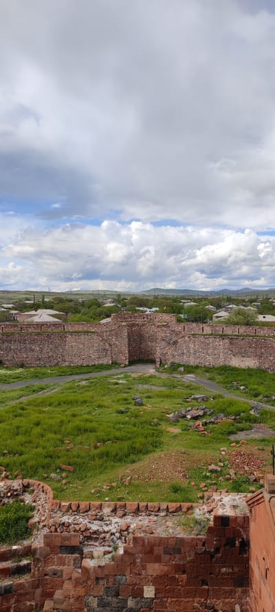 The image presents a view of the Dashtadem Fortress in Armenia, characterized by its aged stone architecture. The foreground features remnants of a brick structure, likely part of the fortress's walls, showing signs of wear and decay. Beyond this, a green field stretches towards a substantial stone wall, which forms a central element of the fortress. The wall is constructed from roughly hewn stones and has an opening leading into the fortress's interior. In the distance, residential buildings are visible, providing a contrast between the historical structure and the contemporary surroundings of Dashtadem. The sky is overcast with scattered clouds, adding to the somber atmosphere of the scene.