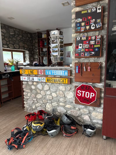 The scene captures the interior of a business, likely a shop or tourist center in Pejë, Kosovo, as indicated by the signage on display. The walls are made of stacked stones. Various items related to tourism and outdoor activities are exhibited. These include magnets, keychains, and signs representing Kosovo and Albania, with specific mentions of "Peja," "Zip Line," "Via Ferrata," and "Marimangat." A sign boldly states, "Never Stop Climbing." Climbing helmets and harnesses are arranged on the floor, indicating that the establishment may be associated with climbing or adventure sports. A display rack features photographs of what appears to be local scenery. A window provides natural light, revealing indoor plants. Shelving units house additional items and equipment.