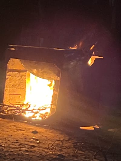 The image depicts a night scene with a large, open structure that appears to be a kiln or furnace. Intense orange and yellow flames are visible erupting from within the structure, illuminating the rough, stone-like interior walls. Branches and twigs are scattered around the base of the structure, some appearing to be fuel for the fire. The ground is rough and uneven, covered in what looks like dirt and debris. In the upper right quadrant of the image, a small, detached object, possibly a piece of burning material, is seen floating in the air against the dark background. The overall atmosphere is dark and smoky, with the fire being the primary source of light. The scene is located in Balkh, Tajikistan.