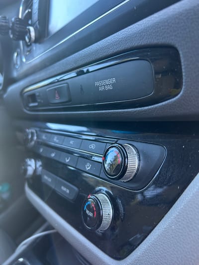 The image shows a close-up of a car's climate control panel. There are two rotating dials with segmented rings of red and blue to indicate temperature settings. Above the dials, there are buttons with various icons and text labels, including "PASSENGER AIR BAG" and a hazard light symbol. The panel is made of black plastic with a glossy finish, and some dust is visible on the surface. The car appears to be parked, with a blurred background that suggests an outdoor environment, possibly a parking lot or street. The lighting suggests it is daytime. The overall impression is that of an interior car shot focusing on the dashboard controls. The location in the city of Everett, United States of America, is not directly discernible from the visual information in the image.