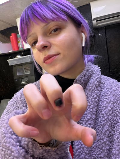 The image depicts an indoor scene in Butsenino, Russia. A person with purple-dyed hair and bangs, wearing a light purple fleece jacket over a black high-necked shirt, is visible from the chest up, looking directly at the camera. A white wireless earbud is present in the individual's right ear. The right hand is extended into the foreground, with fingers curled into a claw-like posture; the thumbnail appears dark or black. The background to the right features a dark wall with a white rectangular air conditioning unit, branded "Daewoo," mounted on it. To the left, a counter or shelving unit displays red and white rolled items, several bottles, a clear plastic storage bin, and a black appliance with a digital display and "iCeon" branding. A gray upholstered surface is partially visible in the lower-left corner. The scene is illuminated by artificial light.