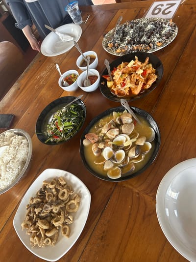 A spread of Indonesian food is displayed on a wooden table. In the foreground, a white oval plate holds a serving of fried calamari rings. To the left, a large metal bowl contains steamed white rice. To the right of the calamari, a black oval bowl is filled with clams in a light yellow broth, garnished with herbs and sliced chilies. Further back, a black oval bowl contains stir-fried water spinach with chili and bean sprouts. A red-chili sauce is served in a small white bowl, alongside another white bowl holding a mixed vegetable relish. A white plate, possibly used or waiting to be served, is positioned at the far right. In the background, a rectangular platter is covered with fried bananas drizzled with chocolate sauce and grated cheese. A table number sign displaying "29" is propped up behind the platter. A person's arm, wearing a dark blue long-sleeved shirt, is visible on the left, holding a white plate. A partially filled plastic cup with a blue lid sits on the table. The scene is indoors, likely in a restaurant.